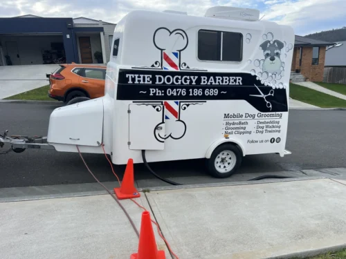 A white mobile dog grooming trailer labeled "The Doggy Barber" is parked on a residential street. The trailer features dog graphics and contact information, and is connected to an orange SUV. Orange traffic cones are placed around the trailer.